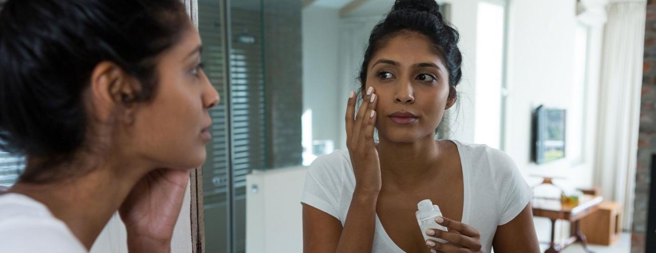 A woman applies sunscreen to her face in front of a mirror